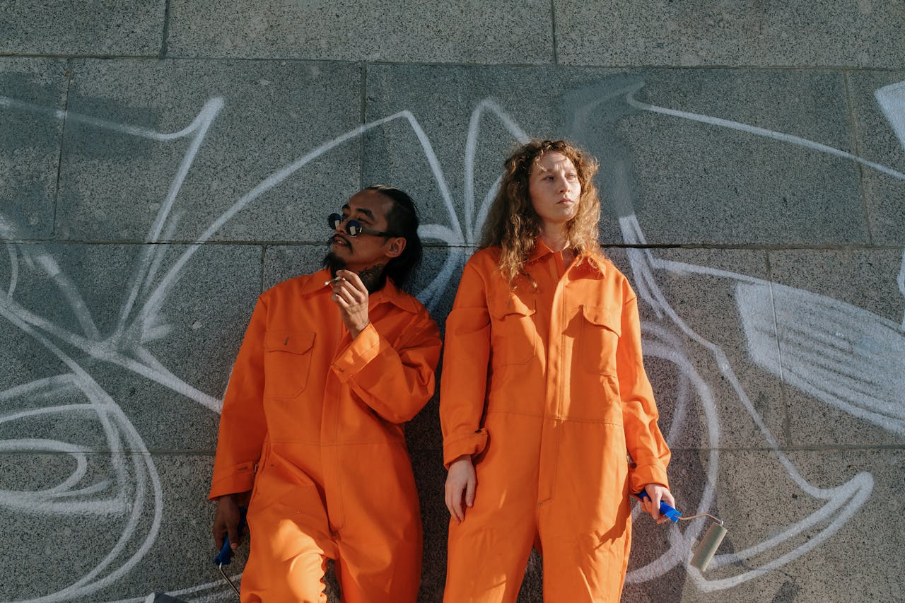 Two community workers in orange uniforms standing against a graffiti wall, holding paint rollers outdoors.