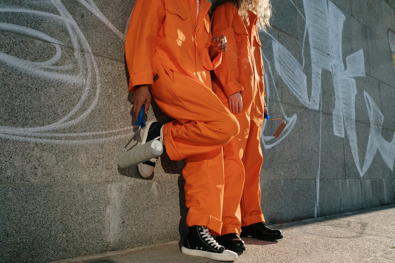 Two people in orange uniforms with paint rollers leaning on a graffiti-covered wall outdoors.