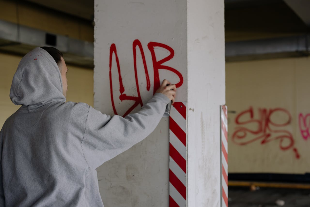 Hooded artist spray painting graffiti on a concrete wall in an urban setting.