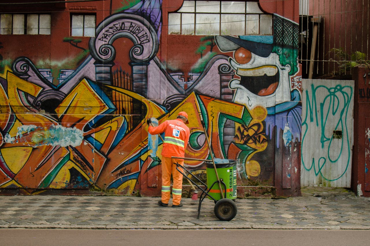 Urban street cleaner in orange uniform working against a vibrant graffiti wall.